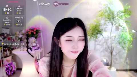 Media: Video of an Asian woman with long black hair, wearing a light pink top, smiling in a bright, sunlit room with floral decorations and a large green plant in the background.