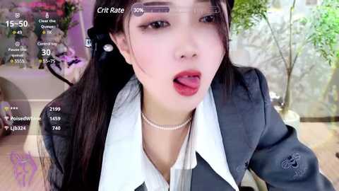 Media: Video of a young Asian woman with fair skin, wearing a gray blazer and white blouse, and making a suggestive face. Background shows a blurred indoor setting with plants.