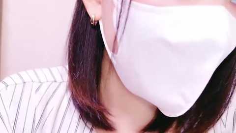 Media: A close-up video of a person with long, straight dark hair, lifting a white shirt to reveal a white bra. The background is a blurred, light-colored wall.