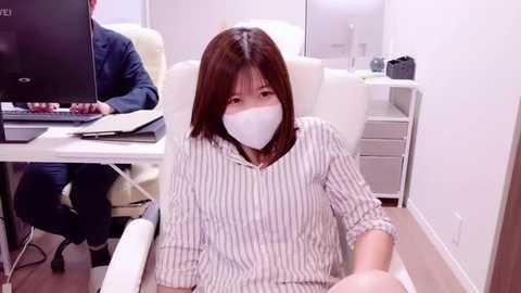 Media: Video of an Asian woman in a white striped shirt and face mask, sitting in a modern office with white furniture and computers, wearing a blue jacket.