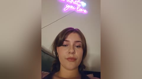 Media: Video of a young woman with fair skin, brown hair, and subtle makeup, wearing a black top. She has a serene expression, with a neon sign above her reading \"I love you.\