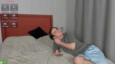 Media: Video of a young man with short brown hair, lying on a bed, wearing a gray shirt and light blue shorts. Background features a red headboard and gray curtains.