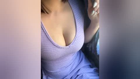 Media: Video of a woman with fair skin, wearing a low-cut, ribbed purple dress, revealing ample cleavage. She has a manicured hand with white nails, holding a smartphone, seated in a car.