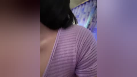 Media: A close-up video of a woman with light skin, black hair tied back, wearing a ribbed, long-sleeve, light purple top. Background features a patterned wall and a blurred person.