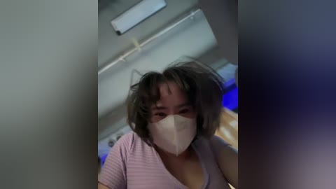 Media: Video of a woman with short, messy brown hair and a beige surgical mask, wearing a light pink short-sleeve shirt, captured from a low angle in a dimly lit room with a white ceiling and blurred background.