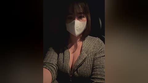 Media: Video of a woman with a white face mask and brown hair, wearing a grey zip-up sweater, seated in a dimly lit room.