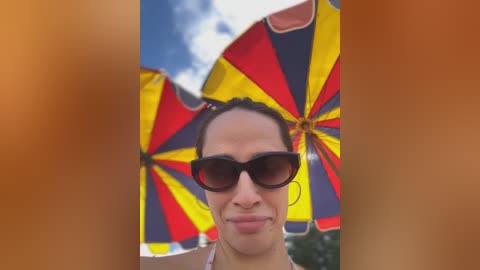 Media: A video of a woman with dark skin, wearing black sunglasses and a red, yellow, and blue umbrella. She has a wide smile and a neutral background of a bright sky and a blurred brown object.