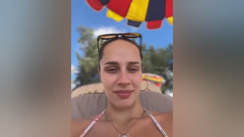 Media: Video of a woman with olive skin, brown hair, and glasses, wearing a colorful bikini, sitting under a beach umbrella.