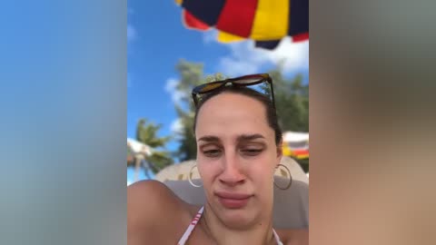 Media: Video of a fair-skinned woman with brown hair, wearing sunglasses and a pink bikini, sitting on a beach chair under a colorful umbrella.