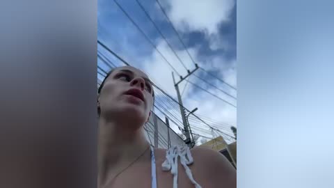 Media: Video of a woman in a white bikini, looking up at a cloudy sky, with power lines and buildings in the background.