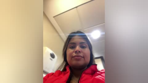 Media: Video of a young woman with medium-length dark hair, wearing a red puffer jacket, standing in a bathroom with beige walls, a white dispenser, and fluorescent lights.