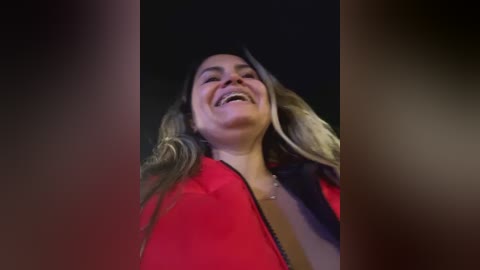 Media: Video of a joyful woman with long, wavy blonde hair, wearing a red jacket, laughing with her mouth open, against a dark background.