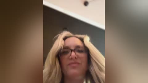 Media: A video of a blonde woman with glasses, wearing a light-colored top, captured from a low angle, emphasizing her hair and face.