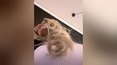 Media: Video of a woman lying on her back, her head tilted upwards, with long, wavy blonde hair cascading down. She wears a white ribbed top and has a serene expression. Background shows a white ceiling and black wall.