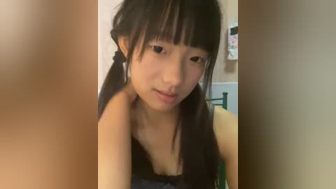 Media: A video of an Asian woman with long black hair in pigtails, wearing a black tank top, captured in a dimly lit room.