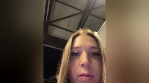 Media: Video of a young Caucasian woman with light skin, long blonde hair, and dark eyebrows, looking directly into the camera, taken indoors with a dim, industrial ceiling featuring metal gridwork.