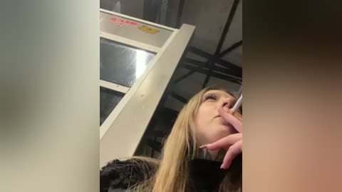 Media: Video of a blonde woman with long hair, wearing a black coat, looking contemplative on a subway platform with metal rails and glass windows in the background.