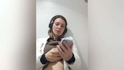 Media: A video of a woman with fair skin, dark hair, wearing headphones, a brown sweater, and a white jacket, taking a selfie in a white-walled, narrow space.