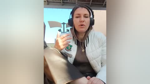 Media: Video of a white woman with long brown hair wearing a white puffer jacket, black headphones, and a gray sweater, sitting on a couch, holding a fan, in a modern living room.