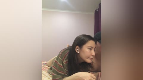 Media: Video of a young Asian woman with long black hair, wearing striped pajamas, lying on a bed with a patterned blanket, listening to earbuds, and kissing a man's leg.