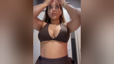 Media: Video of a curvy woman with medium skin tone, medium-length brown hair, and large breasts, wearing a black sports bra and black high-waisted shorts. She stands indoors, adjusting her hair, with a blurred background.