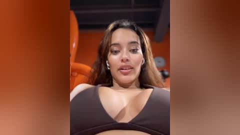 Media: A video of a young woman with light brown skin and long, straight hair, wearing a black bra, seated in a vibrant orange chair, indoors.