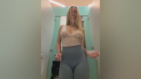 Media: A video of a smiling blonde woman with fair skin, wearing a lacy pink bralette and high-waisted blue jeans, standing in a narrow hallway with mint green walls and a white door.