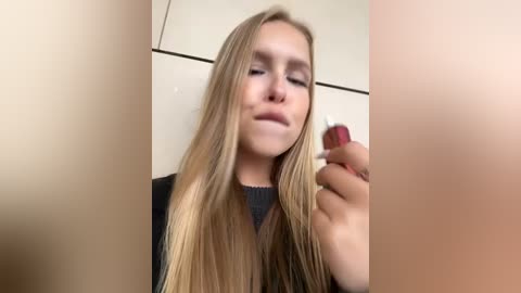 Media: Video of a young Caucasian woman with long, straight blonde hair, light skin, and closed eyes, holding a red lipstick in her right hand. She wears a black top. Background is a beige wall.