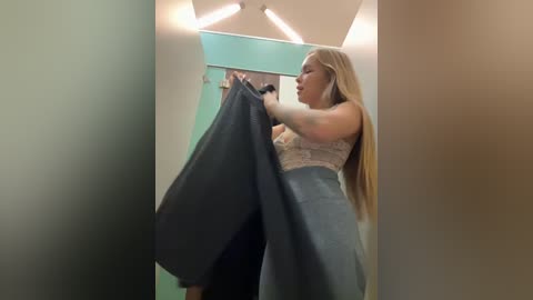 Media: Video of a blonde woman with long hair, wearing a lace bra and a gray dress, adjusting her dress in a dimly lit dressing room with beige walls.