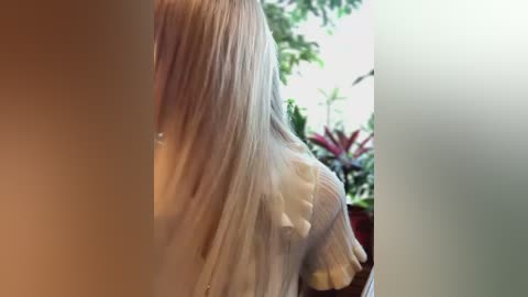 Media: A video showing the side profile of a person with long, straight, platinum blonde hair, wearing a white, sheer, long-sleeved top. The background features a blurred, green, leafy plant and a white wall, creating a serene, indoor setting.