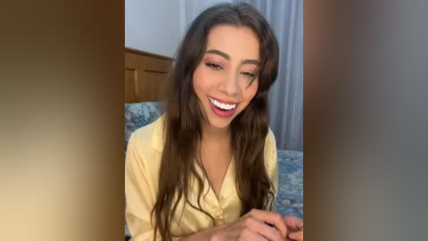 Media: Video of a smiling woman with long brown hair, wearing a yellow shirt, sitting on a bed with patterned blue and white sheets.