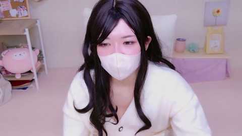 Media: Video of an East Asian woman with long black hair, wearing a white face mask and a cream-colored cardigan, sitting on a light beige carpet in a pastel-colored room with soft toys and framed artwork in the background.