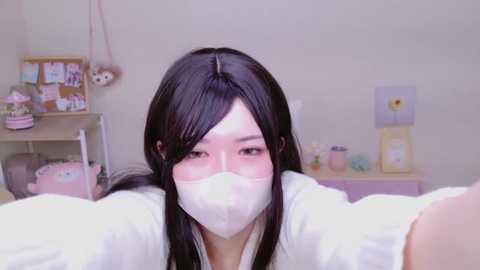 Media: Video of an Asian woman with long black hair and light skin, wearing a white face mask and white jacket, taking a selfie in a pastel-themed room with a bulletin board, flower decorations, and pastel-colored furniture.