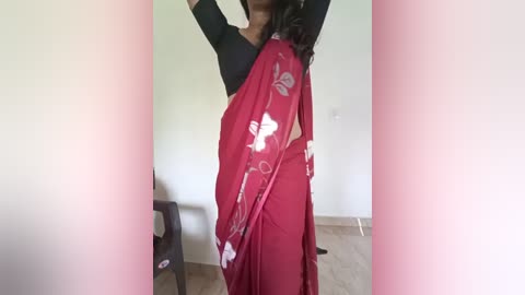 Media: Video of a woman in a red sari with white floral design, wearing a black top, standing in a simple room with beige floor.