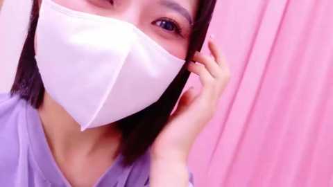 Media: Video of an East Asian woman with straight black hair, wearing a white surgical mask, light purple scrubs, and pink curtains in the background.