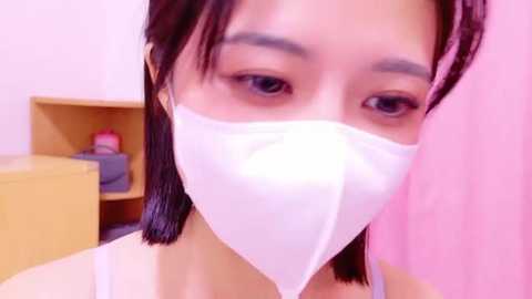 Media: A close-up video of an Asian woman with medium skin tone, wearing a white face mask, looking directly at the camera with brown eyes and straight black hair. Background features a pink wall and a wooden shelf with a red item.