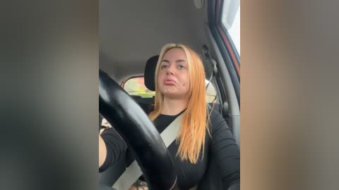 Media: A video of a blonde woman with long hair, wearing a black top, driving a car with a black steering wheel, seen through a car window.