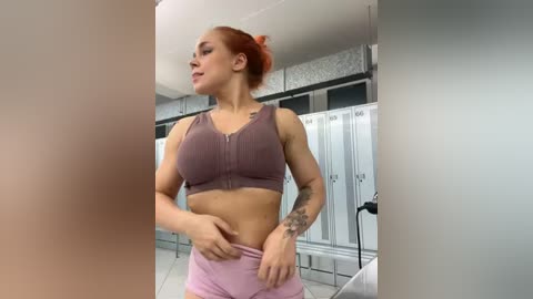 Media: Video of a fit woman with red hair tied back, wearing a purple sports bra and pink shorts, posing in a gym locker room with white lockers and fluorescent lighting.