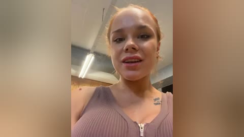 Media: A video of a young woman with light brown skin and short hair in a ponytail, wearing a ribbed, sleeveless top. She has a small tattoo on her upper chest. The background shows an indoor setting with fluorescent lighting and exposed ceiling.