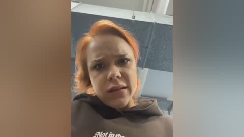 Media: Video of a woman with short, bright orange hair and pale skin, wearing a dark brown hoodie, looking slightly confused or surprised. Background includes a grey and white tiled wall.