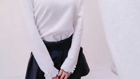 Media: Video of a person with light skin wearing a white long-sleeved blouse and a black skirt with lace trim, standing against a plain white background.