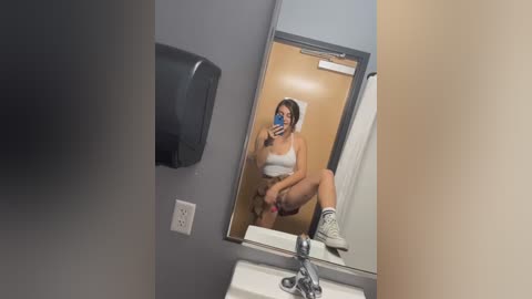 Media: Video of a young woman in a public restroom mirror, wearing a white sports bra and white socks, holding a phone, exposing her genitals, in a casual, unflattering pose.