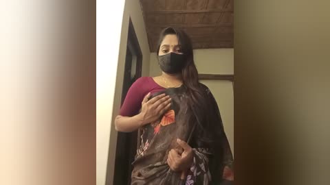 Media: Video of a South Asian woman with medium skin tone, wearing a maroon top and black mask, holding a black sari with floral design, in a dimly lit room with wooden ceiling.