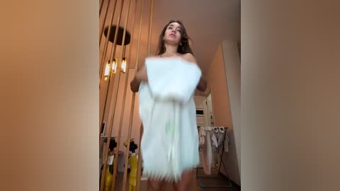 Media: Video of a young woman with long brown hair, wearing a white fluffy dress, standing in a dimly lit room with a beige wall and hanging plants, holding a fluffy pillow.