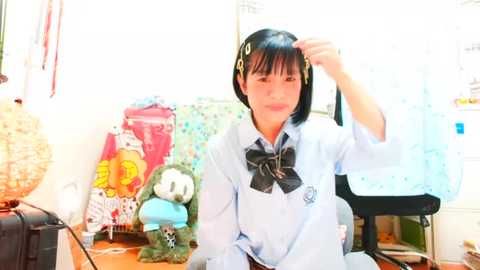 Media: Video of a young Asian woman with short black hair, wearing a light blue school uniform with a black bow, adjusting her hair in a cluttered room filled with stuffed toys and decorations.