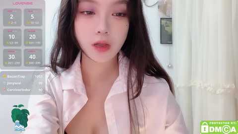 Media: A video of an Asian woman with long black hair, wearing an unbuttoned white shirt, revealing cleavage, standing in a brightly lit room with a calendar and green curtain in the background.