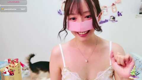 Media: Video of a smiling young Asian woman with dark hair, wearing a white lace bra, playfully covering her mouth with a pink bandana in a cozy bedroom with a cat and wall decorations.