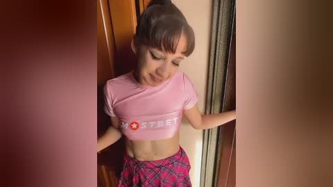 Media: Video of a young woman with light skin and brown hair in a ponytail, wearing a pink cropped top and plaid skirt, leaning against a wooden door.