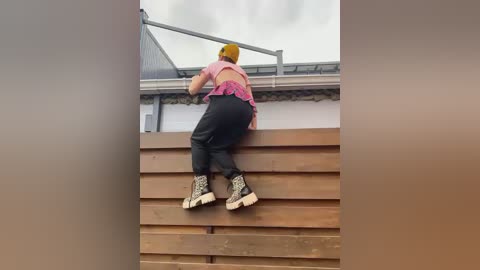 Media: Video of a person in a yellow helmet climbing wooden steps on a rooftop, wearing a pink tank top and black pants, under a cloudy sky.