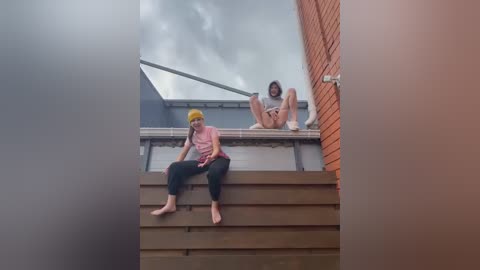 Media: A video of two women on a rooftop, one in a pink top and yellow beanie, the other in a gray hoodie and shorts, against a cloudy sky and brick wall.